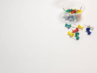 Renkli bir itme iğnesi koleksiyonu, kırmızı, yeşil, mavi, sarı ve beyaz, temiz beyaz bir zemin üzerine saçılmıştır. Bazı iğneler şeffaf plastik bir kabın içinde gruplandırılırken, diğerleri ayrı olarak düzenlenir. Resim yeterli sayıda kopya sağlar