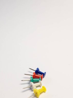 Sade beyaz arka planda köşegen olarak dizilmiş beş renkli raptiyeli yakın çekim bir stüdyo resmi. İtme iğneleri mavi, kırmızı, yeşil, beyaz ve sarıdır. Bileşimin, iğnelerin başı ve iğnelerine odaklanan sığ bir alan derinliği var. Lütfen!
