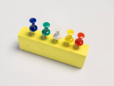 Yakın plan bir stüdyo çekiminde, canlı sarı dikdörtgen bir blokta düzgünce hizalanmış beş renkli raptiye yer alıyor. İğneler art arda diziliyor, her birinin kafası farklı renklerde, görsel olarak çekici ve düzenli bir görüntü yaratıyor. Arka plan bir...