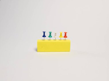 Basit bir stüdyo çekiminde üzerinde beş farklı renk iğnesi bulunan sarı dikdörtgen bir blok bulunur. İğneler sıraya dizildi. Arkaplan bembeyaz, temiz ve minimalist bir kompozisyon oluşturuyor. Görüntü iyi aydınlatılmış ve...