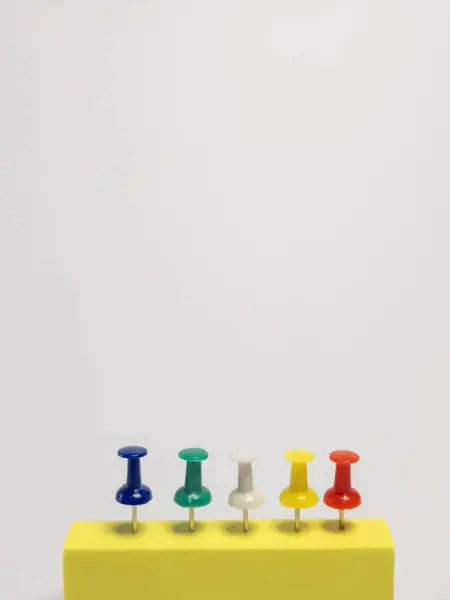 Dikey bir stüdyo çekiminde, geniş bir kopya alanı olan düz beyaz bir arkaplan üzerine yerleştirilmiş, sarı dikdörtgen bir bloğa yerleştirilmiş beş renkli raptiye sırasına yer verilir. İtme iğneleri mavi, yeşil, beyaz, sarı ve kırmızıdır. Metal iğneleri temizdir.