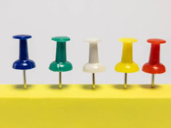 Yakın çekim stüdyosunda beş farklı renk iğnesi üst üste düzgünce yerleştirilmiş. Çiviler sarı bir yüzeye gömülmüş ve beyaz arkaplana karşı canlı bir kontrast oluşturuyor. Resim, günlük nesnenin basit güzelliğini vurgular.