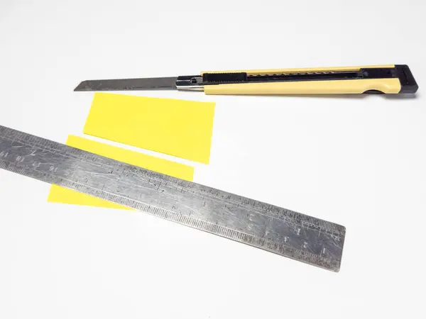 Beyaz bir yüzey üzerinde tepeden tırnağa bıçak, metal cetvel ve sarı kağıt. Kağıt kısmen kesilmiş, bu da bir işçilik ya da kesim sürecini gösteriyor. Bileşim kesinliği ve ölçüm ve kesim için kullanılan araçları vurguluyor. Görüntü