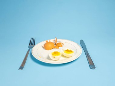 Bir natürmort fotoğrafında iki yarısı haşlanmış yumurta ve yumurta kabuğu olan beyaz bir tabak bulunur. Açık mavi bir yüzeye bir çatal ve bıçak yerleştirilir. Resim parlak bir şekilde ışıklandırılmış, yiyeceklerin ve çatal bıçakların dokularını ve renklerini vurguluyor..