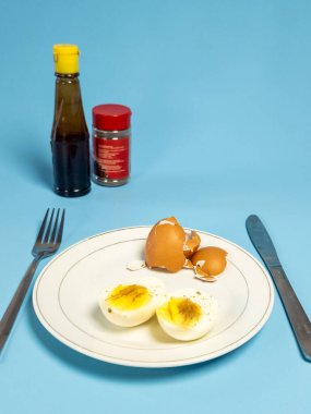 İki parça haşlanmış yumurta, yumurta kabuğu ve çatal ve bıçakla görsel olarak çekici bir fotoğraf. Görüntü bir şişe soya sosu ve açık mavi arka planda bir kutu biber içeriyor. Odak noktası yemek ve basit halkla ilişkiler.
