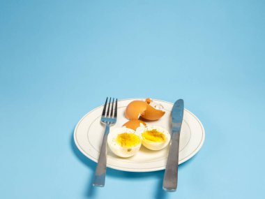 Bir çatal ve bıçakla birlikte beyaz bir tabakta sunulan yarı haşlanmış bir yumurtayı içeren bir fotoğraf. Sahne, temiz ve minimalist bir estetik yaratmak için mavi bir zemine dayanır. Yumurta kabukları da mevcuttur. Kompozisyon s