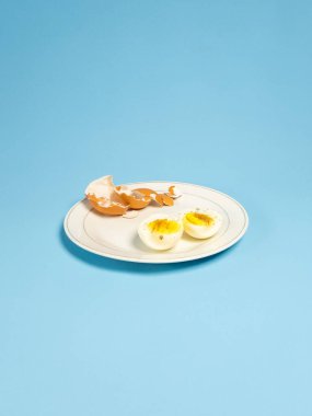 Haşlanmış yumurta ve yumurta parçalarıyla dolu beyaz bir tabağın yer aldığı bir stüdyo resmi. Tabak açık mavi bir arka planda duruyor. Yumurtalar biberle tatlandırılmış ve taze hazırlanmış görünüyor. Görüntü basit, temiz ve uygun. 