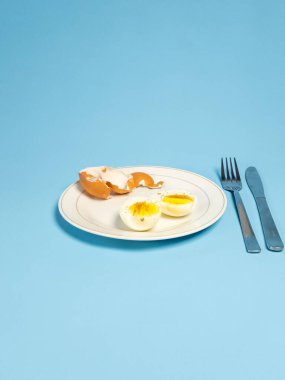 Yukardan aşağıya doğru iki yarısı haşlanmış yumurta, yumurta kabuğu, çatal ve açık mavi arka planda bir bıçak olan bir tabağı gösteriyor. Yumurtalar biberle tatlandırılmış. Görüntü basit, temiz bir estetik, besinle ilgili projeler için mükemmel.