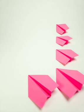 Yukarıdan aşağıya doğru çekimde düz beyaz bir yüzey üzerinde çapraz çizgi halinde dizilmiş bir dizi pembe kağıt uçak bulunur. Görüntü, origami uçaklarının şekillerini ve canlı renkle temiz zemin arasındaki zıtlığı vurguluyor. Kimlik