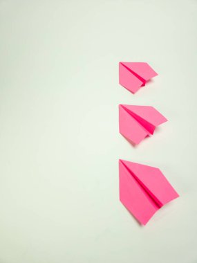 Bir stüdyo çekiminde, temiz beyaz arka planda çapraz dizilmiş üç pembe kağıt uçak yer alır. Görüntü, kompozisyonun basitliğini ve kağıt uçakların canlı rengini vurguluyor. Uçaklar eşit aralıklarla bir his yaratıyor.