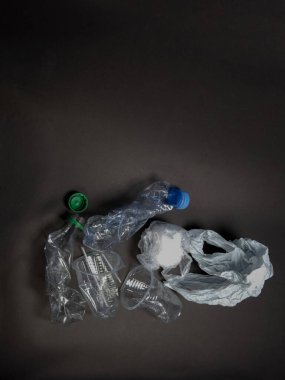 Şişeler, fincanlar ve koyu gri bir yüzeye saçılmış bir poşet de dahil olmak üzere çeşitli buruşuk plastik parçaların yukarıdan aşağı görüntüsü. Görüntü, plastik atık sorununu ve çevre üzerindeki etkisini vurguluyor. Kompozisyon basit, Vol 'u vurguluyor.