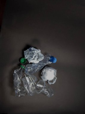 Ezilmiş plastik şişeler, fincanlar ve koyu gri bir yüzey üzerinde buruşuk kağıtlar. Kompozisyonda plastik atık ve çevre kirliliği sorunu vurgulanıyor. Işıklandırma yumuşak, gölgeler görüntüye derinlik ve doku katıyor.. 