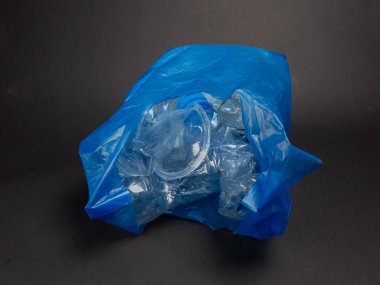 Yakın plan çekimde, şeffaf plastik şişeler ve kaplarla dolu buruşuk mavi bir plastik torba görülüyor. Poşet koyu gri bir yüzeyde duruyor ve içindekiler görünür durumda, atık, geri dönüşüm ve çevresel kaygılar kavramını vurguluyor. Şey... 