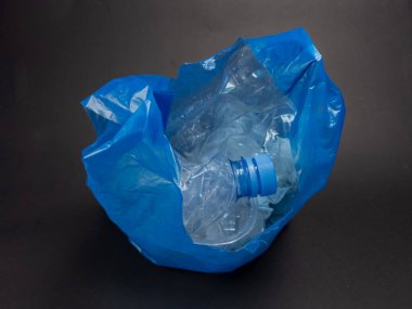 Buruşmuş plastik şişeler ve atıklarla dolu mavi plastik bir torba, karanlık bir arkaplana kurulmuş. Görüntü, plastik kirliliği ve çevre üzerindeki etkisini vurguluyor. Odaklandığımız nokta iki nokta arasındaki görsel zıtlık.