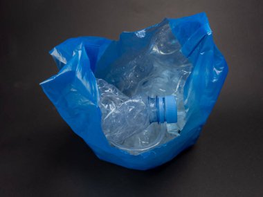 Yakın plan çekimde, kullanılmış ve ezilmiş plastik şişelerle dolu buruşuk mavi bir plastik torba görülüyor. Poşet açık, içindeki şişeleri ortaya çıkarıyor, bir şişe de bariz bir şekilde sergileniyor. Arkaplan, atığı vurgulayan sade bir karanlık yüzey.