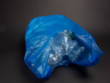 Buruşmuş, berrak plastik şişelerle dolu mavi bir plastik torba koyu gri bir arka planda duruyor. Görüntü, plastik atık ve geri dönüşüm sorununu vurguluyor. Işıklandırma yumuşak ve doğal, plastiğin dokularını ve şekillerini vurguluyor. T