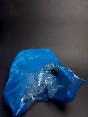 Yakın çekim, buruşmuş, şeffaf plastik şişelerle dolu mavi bir poşet yakalıyor. Koyu gri arka planda duran çanta, atıkları ve geri dönüşüme duyulan ihtiyacı vurguluyor. Kompozisyon plastiğin dokusunu vurguluyor.