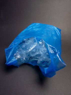 Mavi bir plastik torba buruşuk, şeffaf plastik şişelerle dolu, muhtemelen geri dönüşüm için atılıyor. Görüntü koyu gri bir arkaplanda çekiliyor ve parlak mavi çanta ile açık plastik arasındaki zıtlığı vurguluyor. Kompozisyon yüksekliği