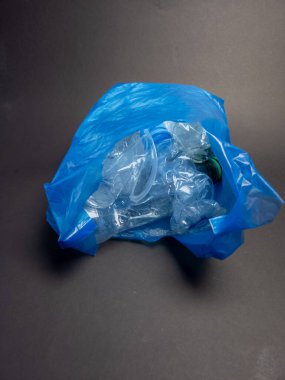 Plastik atık ve kirlilik sorununu gösteren çeşitli ezilmiş ve atılmış plastik şişe ve konteynırlarla dolu mavi bir plastik torba. Resim koyu gri bir arkaplan üzerinde çekilir, mavi ba arasındaki kontrastı vurgular