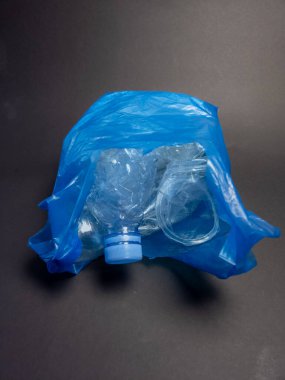Plastik şişeler ve diğer plastik atıklarla dolu mavi bir poşetin yakın plan çekimi, plastik kirlilik ve çevresel kaygıları vurguluyor. Resim koyu gri bir arkaplanda çekiliyor, konuyu ve kreasyonu vurguluyor.
