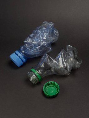 Esmer arka planda atılmış ve ezilmiş iki plastik su şişesi. Bir şişenin mavi bir kapağı, diğerinin yeşil bir kapağı var ve yeşil kapak ayrı ayrı yatıyor. Görüntü atık, geri dönüşüm ve çevresel sorunların temalarını aktararak,