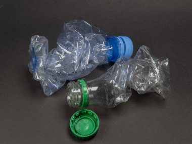 Koyu gri bir yüzeye iki plastik şişe ve iki şişe kapağı yakın çekim. Şişeler şeffaf, kullanıldıktan sonra şekillerinin bozulduğunu gösteriyor. Bir şişenin mavi kapağı, diğerinin yeşil kapağı var. Bu görüntü çevreyi temsil ediyor 