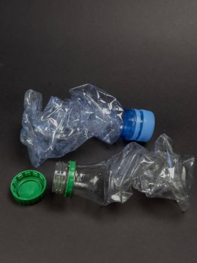 İçinde iki ezilmiş plastik su şişesi ve karanlık arka planda atılmış bir kapağın olduğu bir fotoğraf. Şişeler buruşmuş ve deforme olmuş ve plastik atık sorunu ve bunun çevre üzerindeki etkisini vurguluyor. Kompozisyon vurgulanıyor.
