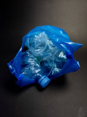 Yakın plan çekimde, muhtemelen geri dönüşüme gidecek olan buruşmuş plastik şişe ve bardaklarla dolu mavi bir plastik torba görülüyor. Görüntü, plastik atık sorununu ve çevreyi korumanın önemini vurguluyor. Koyu arkaplan karşıtlığı