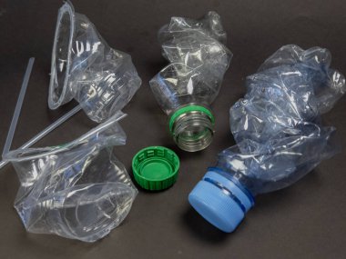 Yakın çekimde, ıskartaya çıkarılmış plastik şişeler ve şişe kapakları koyu bir zemine saçılmış. Görüntü, plastik atıkları ve çevre üzerindeki etkisini vurguluyor. Şişelerin farklı renkte kapakları var ve kullanıldığına dair belirtiler var.