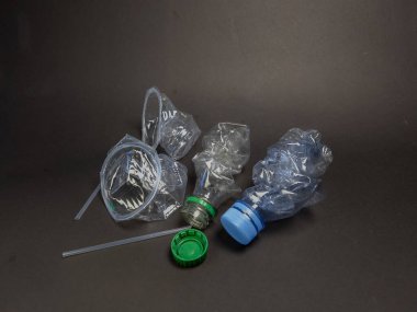 Atılan plastik atık parçalarının yakın plan çekimi. İçinde mavi ve yeşil kapaklı ezilmiş plastik şişeler, yeşil bir kapak ve açık plastik bir pipet var. Atık koyu gri bir arka plana dağılmış durumda, çevresel sorunu vurguluyor.
