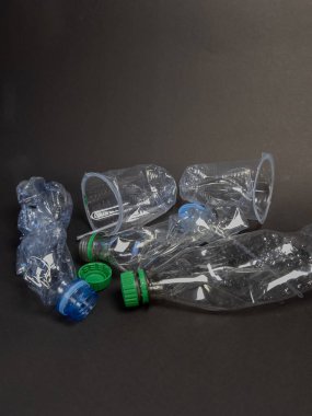 Ezilmiş plastik şişeler ve fincanlar koyu bir zemine saçılmış durumda. Görüntü atık, geri dönüşüm ve çevresel kaygılar kavramını yakalıyor. Yakın plan çekimde atılanların doku ve şekilleri vurgulanır, vurgulanır