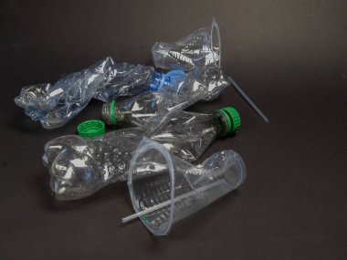 Bu resim, plastik şişeleri ve koyu bir arka plana serpilmiş bir fincanı eziyor. Kompozisyonda, geri dönüşüm ve atıkların azaltılması gereğinin vurgulandığı plastik atık ve çevre kirliliği sorunu vurgulanıyor. Odaklanma metne.