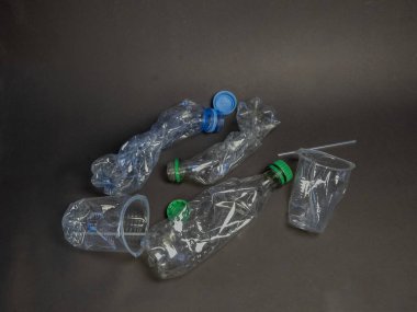 Koyu gri arka plana saçılmış kullanılmış ve ezilmiş plastik şişeler ve bir kupa koleksiyonu. Kompozisyonda plastik atıklar ve bunun çevresel etkileri vurgulanıyor. Görüntü, dolandırıcılıkların sonuçlarını gösteren bir atık ve kirlilik hissi veriyor.