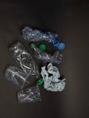 Koyu gri arkaplana saçılmış şişe ve fincanlar da dahil olmak üzere çeşitli ezilmiş plastik atık türlerinin genel görüntüsü. Kompozisyon, plastik kirliliği ve atık yönetimi sorununu vurguluyor. Resim, görüntülerin dokularını ve şekillerini gösterir