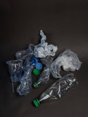 Koyu bir arka planda şişeler, kapaklar, kapaklar ve plastik poşetler de dahil olmak üzere ezilmiş plastik atık koleksiyonu sergileniyor. Görüntü, plastik kirliliği ve geri dönüşüm ihtiyacını vurguluyor. Işıklandırma nötr, metni vurguluyorum.