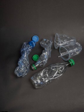 Yüksek açılı çekimde koyu gri bir yüzeye dağılmış ezilmiş plastik şişeler ve bardaklar görülüyor. Görüntü, plastik atık ve çevresel kaygıları vurguluyor. Objeler odak noktasında, karanlık arka plan tarafsız bir arka plan sağlıyor.