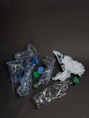 Koyu gri bir yüzeye saçılmış şişe, bardak ve kapak dahil olmak üzere atıImış çeşitli plastik parçaların genel çekimi. Görüntü, plastik atık sorununu ve çevre üzerindeki etkisini vurguluyor. Kompozisyon basit ve net, vurgulu.