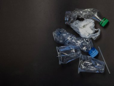 Çöpe atılmış çeşitli plastik şişelerin, bardakların ve bir pipetin yukarıdan aşağıya bakışı. Koyu ve dokulu bir yüzeyde ezilmiş ve buruşmuş. Görüntü, plastik atık sorununu ve çevre üzerindeki etkisini vurguluyor. Kompozisyon w 'nin hacmini vurgular