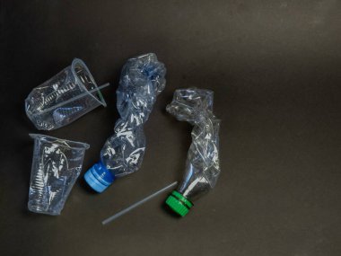 Buruşmuş plastik şişeler, fincanlar ve pipetlerin yer aldığı üstten çekimler koyu bir arkaplanda. Görüntü, plastik atık ve çevre kirliliği sorununu vurguluyor. Kompozisyon ve ışıklandırma, atılan ögelerin dokusunu vurgular