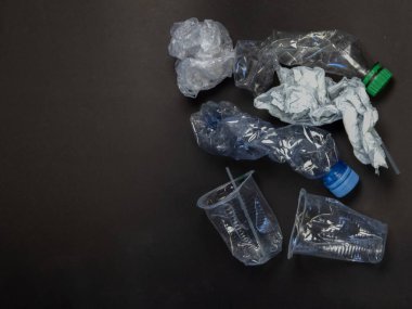 Karanlık bir yüzeye saçılmış şişe ve fincanlar da dahil olmak üzere çeşitli plastik parçaların yer aldığı tepeden tırnağa bir çekim. Kompozisyonda, geri dönüşüm ve atıkların azaltılması gereğinin vurgulandığı plastik atıkların çevresel etkileri vurgulanıyor. Bitti.