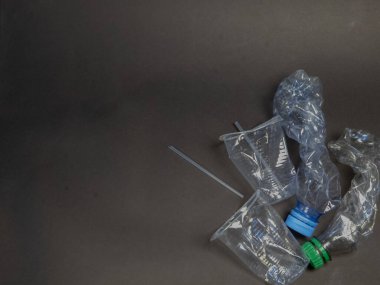 Plastik atık ve kirlilik sorununu vurgulayarak, kırışık plastik şişeler, tek kullanımlık bardaklar ve pipetlerin koyu gri bir yüzeye saçıldığı genel bir çekim. Görüntü, geri dönüşüm çağrısında bulunarak tek kullanımlık plastiklerin çevresel etkilerini vurguluyor.