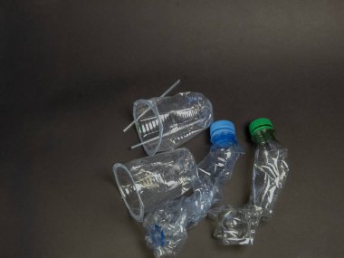 Yukarıdan aşağıya doğru çekimde, koyu gri bir yüzeye saçılmış ezilmiş plastik şişeler ve tek kullanımlık bardaklar görülüyor. Görüntü, plastik atık ve çevre kirliliği sorununu vurguluyor. Kompozisyonda çeşitli bozulmuş plastik kaplar yer alıyor.
