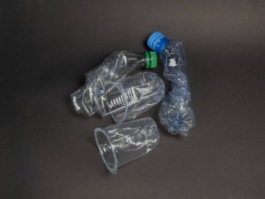Bu resim, plastik atık sorununu vurgulayarak koyu gri bir yüzeye saçılmış çok sayıda buruşmuş plastik şişe ve bardağı gösteriyor. Bileşim, atılan nesnelerin şekillerini ve dokularını vurguluyor. Çevresel farkındalığa odaklanmış durumda.