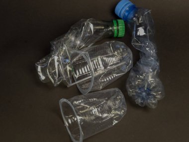 Plastik atık sorununu vurgulayan yassılaştırılmış plastik şişe ve bardaklardan oluşan bir derleme karanlık bir yüzeye uzanıyor. Görüntü buruşmuş formları ve atılanların şeffaflığını gözler önüne seriyor, çevresel kirliliği ve ihtiyacı vurguluyor