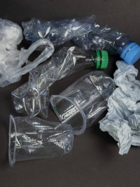 Tepeden çekimde, koyu gri bir yüzeye yerleştirilmiş şişe, bardak ve çantalar da dahil olmak üzere ezilmiş ve atılmış plastik atık koleksiyonu görülüyor. Görüntü çevre kirliliği ve plastik atık sorununu vurguluyor, çeşitli formları gösteriyor