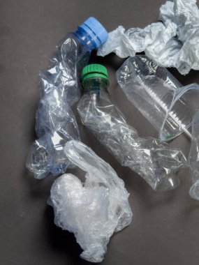 Yüksek açılı çekimde, mavi ve yeşil kapaklı ezilmiş su şişeleri, plastik bardaklar ve buruşuk plastik poşetler de dahil olmak üzere çeşitli plastik eşyalar sergileniyor. Ögeler koyu gri bir yüzeye saçılmış, atık ve çevresel etkenleri vurguluyorlar.