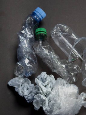 Koyu gri bir yüzey üzerinde buruşmuş plastik şişeler, bardaklar ve plastik poşetler. Görüntü, atık ve çevresel bir endişe hissi veriyor. Atılmış plastiğin dokusu ve şekillerine odaklanarak, pollu sorununu vurguluyor