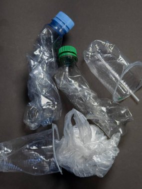 Üzerinde ezilmiş plastik şişeler ve koyu gri bir yüzey üzerinde plastik bir torba olan bir fotoğraf var. Görüntü, plastik atık ve çevre kirliliği sorununu vurguluyor. Kompozisyon basit, dokusu ve şekli o 'ya odaklı.