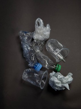 Şişeler, bardaklar ve koyu gri bir yüzeye saçılmış plastik poşet de dahil olmak üzere çeşitli ezilmiş plastik atıkların yer aldığı yukarıdan aşağıya doğru bir çekim. Bu resim plastik atıkların çevresel etkisini vurgular ve geri dönüşüm ihtiyacını vurgular ve 