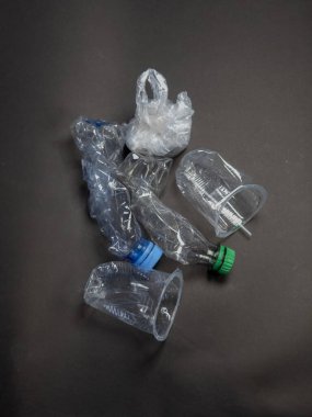 Su şişeleri, bardaklar ve plastik poşet de dahil olmak üzere, atılan plastik atıkların yukarıdan aşağıya doğru bir görüntüsü koyu gri arka planda gösteriliyor. Görüntü, plastik kirliliği ve atık yönetimi sorununu vurguluyor. Odak noktası plastiğin çeşitliliği. 
